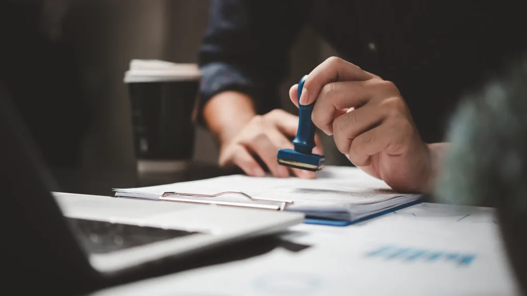 Person stamping documents on a desk with a coffee cup nearby.