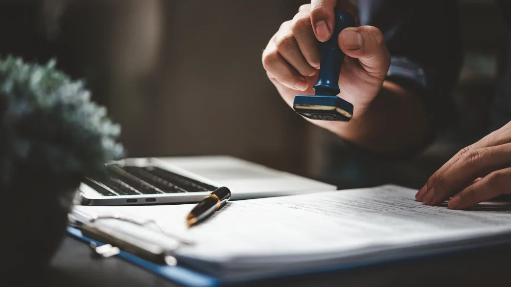 Hand stamping a document on a desk beside a pen and laptop.